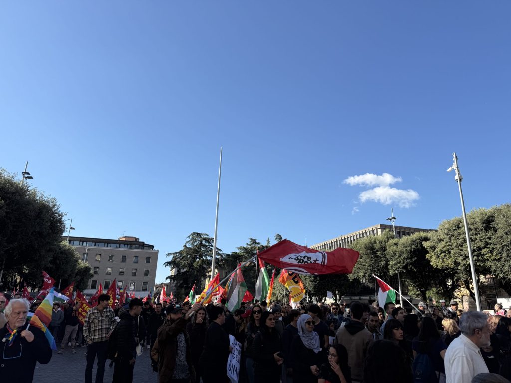Una folla di manifestanti alziamo bandiere e striscioni durante una protesta a Terni, con edifici e alberi sullo sfondo sotto un cielo sereno.