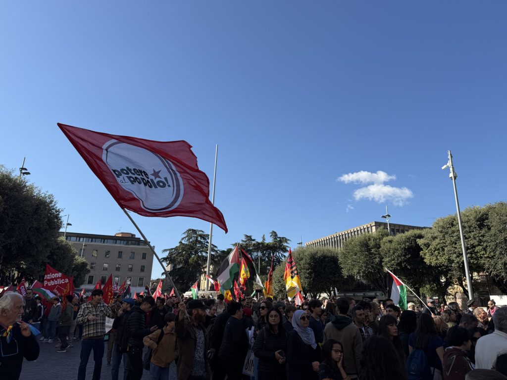 Una folla di manifestanti si raduna in piazza con bandiere e striscioni per una protesta a favore della Palestina, sotto un cielo sereno.