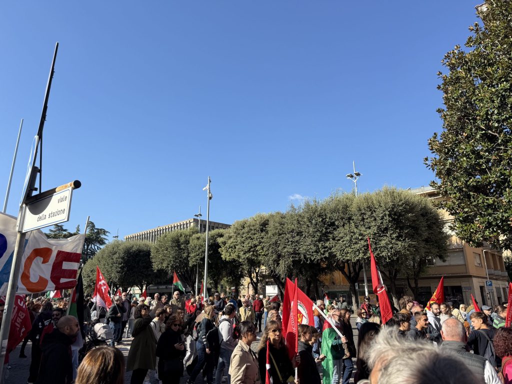 A crowd of protesters with flags gathering in a public space during a demonstration in Terni, under a clear blue sky.