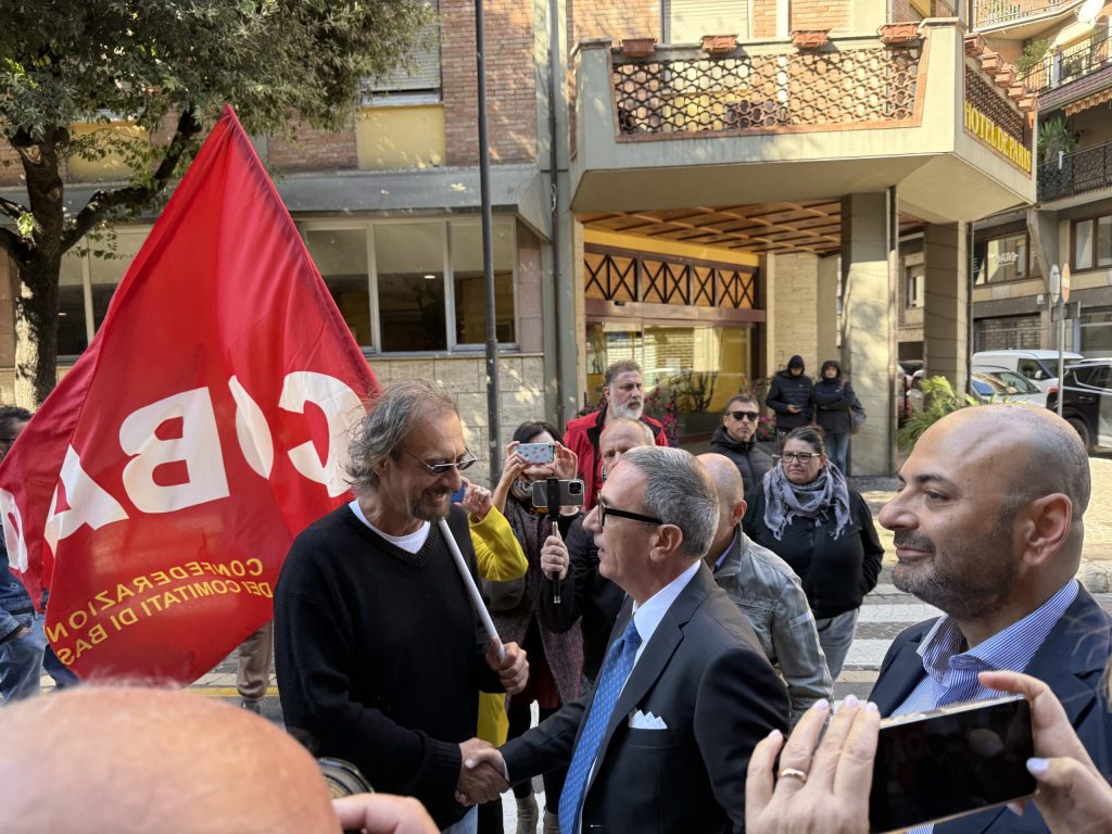 Un gruppo di manifestanti a Terni tiene una bandiera rossa con la scritta 'CGL' mentre un uomo in giacca e cravatta stringe la mano a un uomo con i capelli lunghi, circondati da altre persone che osservano.