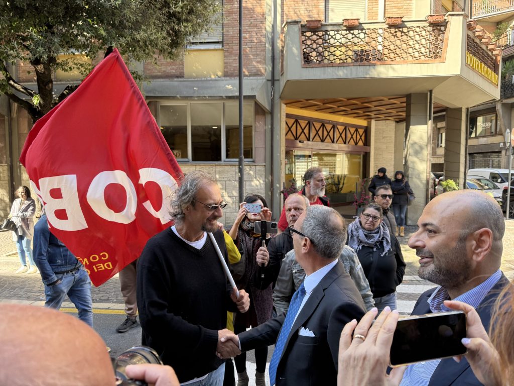 Un manifestante con una bandiera rossa del COBAS stringe la mano a un uomo in giacca e cravatta, mentre altre persone assistono alla scena in un ambiente urbano.