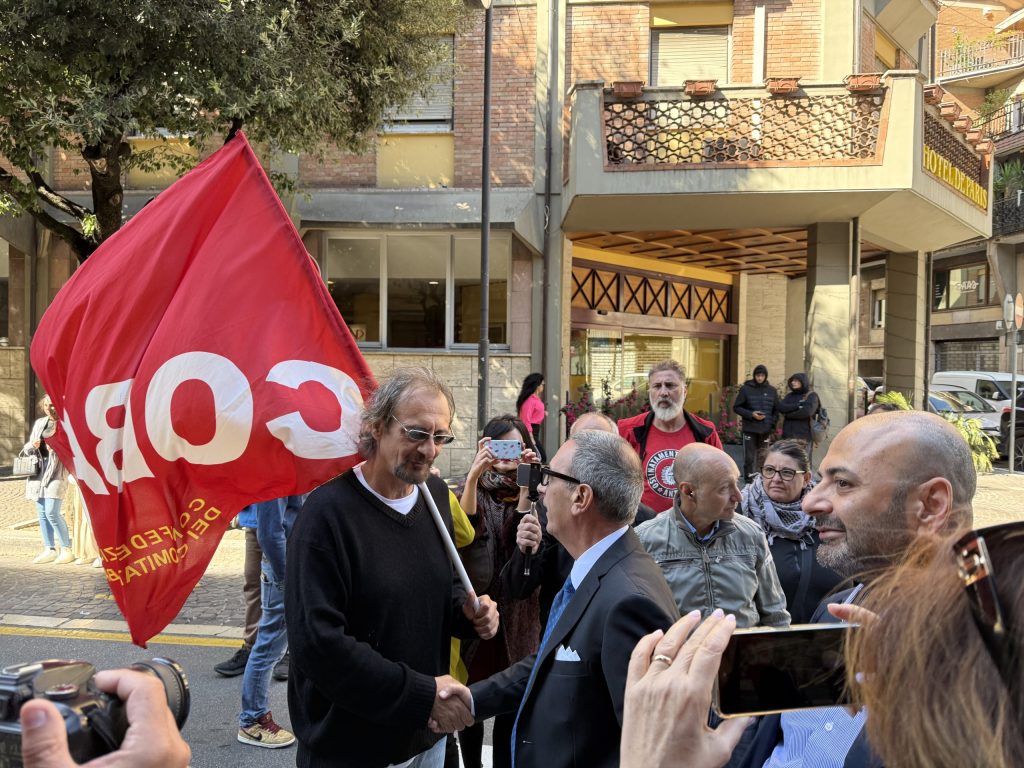 Un uomo con occhiali e capelli lunghi tiene una bandiera rossa con la scritta 'COBAS' mentre stringe la mano a un altro uomo in giacca e cravatta, circondato da una folla di persone che osservano.