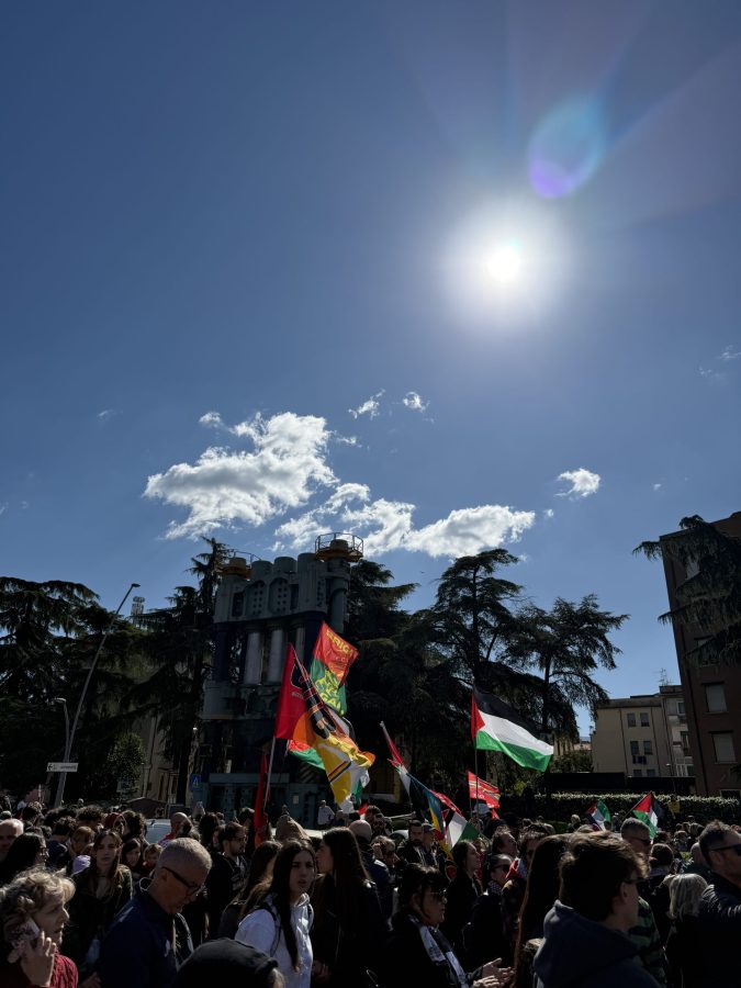 A large crowd of people gathered in Terni for a protest, holding various flags including Palestinian flags, against genocide, with a clear blue sky and bright sun in the background.