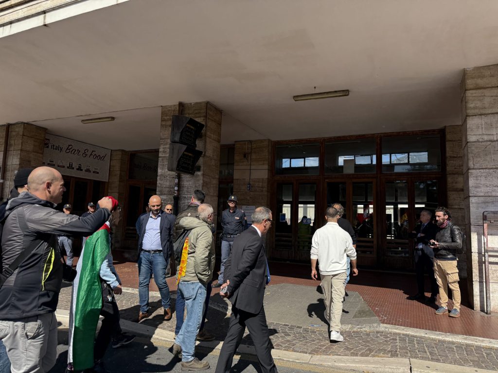 Un gruppo di manifestanti si raduna davanti a un edificio, alcuni indossando abbigliamento colorato, mentre altri interagiscono nel contesto di una manifestazione.