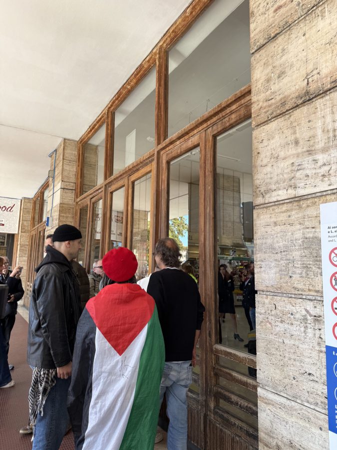 Un gruppo di manifestanti davanti a un edificio, con una persona avvolta in una bandiera palestinese.