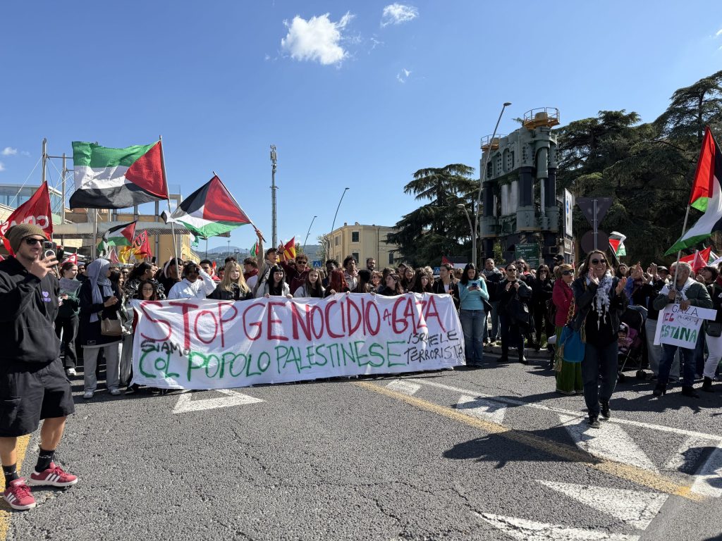 Manifestazione con migliaia di persone che protestano per la Palestina, sventolando bandiere e tenendo striscioni con slogan contro il genocidio.