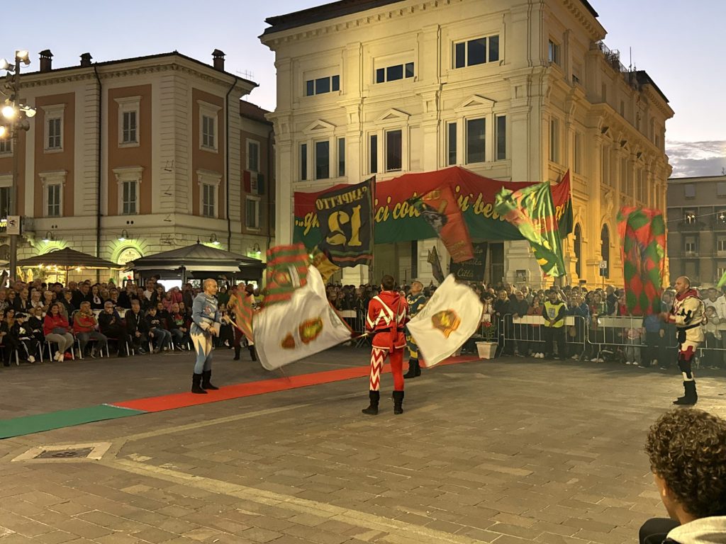 Sbandieratori di San Gemini eseguono una performance in piazza della Repubblica, con un pubblico numeroso e bandiere colorate sullo sfondo.