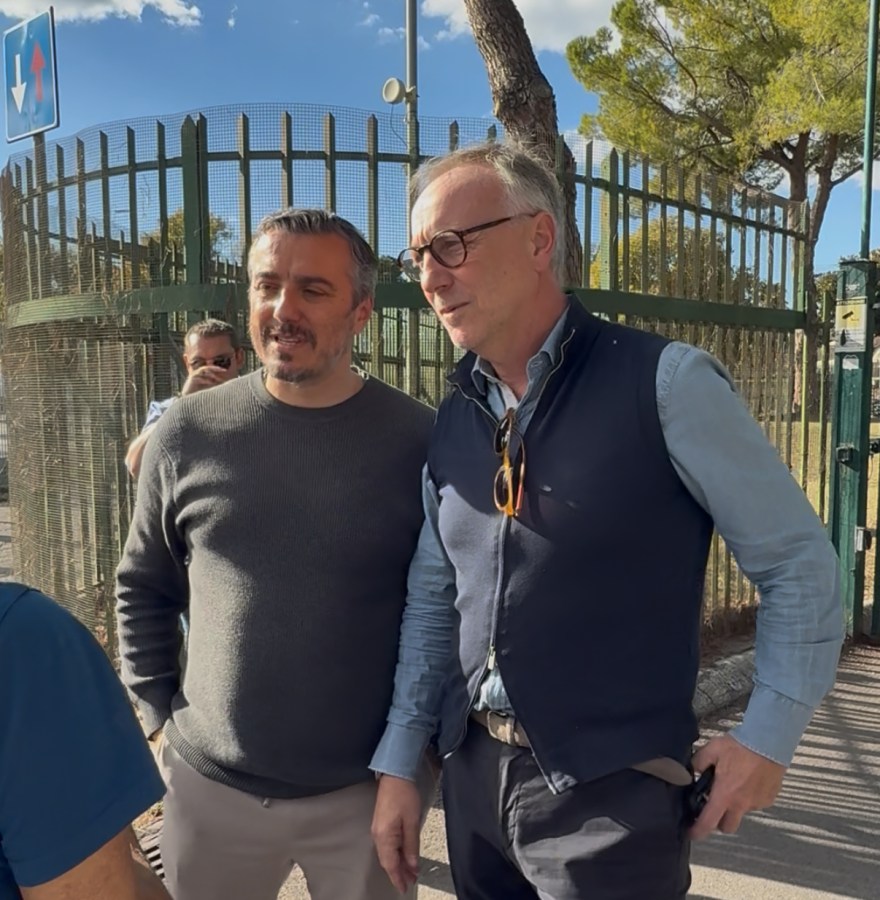 Due uomini sorridenti in un incontro informale presso il cantiere dello stadio a Terni, con recinzione e alberi sullo sfondo.