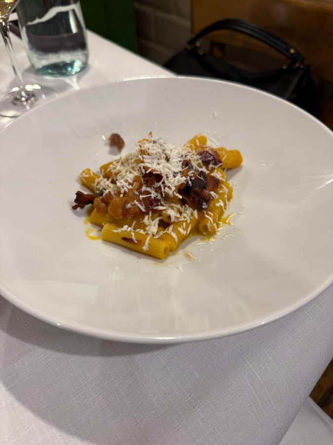 Pasta servita in un piatto bianco, guarnita con formaggio grattugiato e cubetti di pancetta.