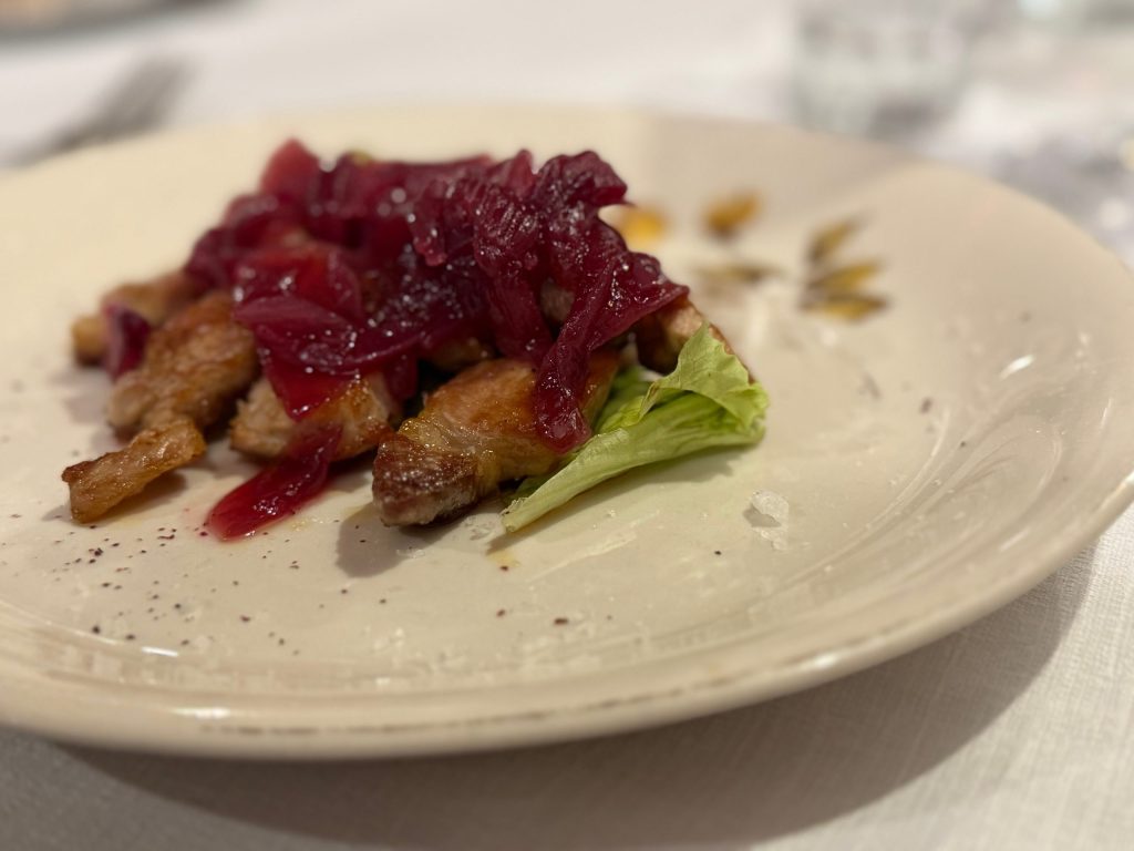 Un piatto gourmet di carne servito su un piatto di ceramica, con strati di cipolle rosse caramellate e una foglia di insalata sul lato.