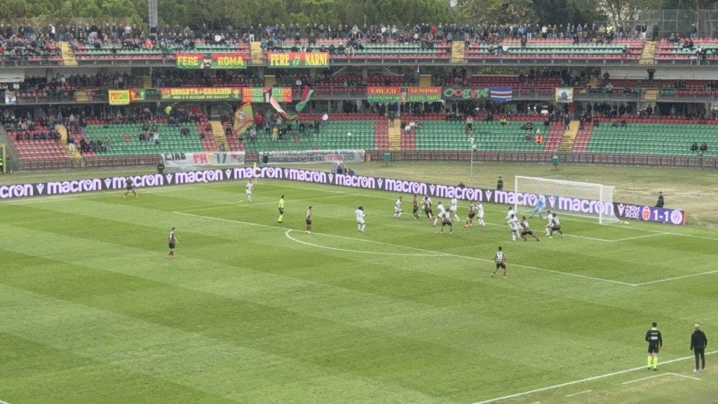Immagine di una partita di calcio in corso, con il campo verde e le tribune piene di tifosi. Si vedono giocatori in azione e pubblicità a bordo campo.
