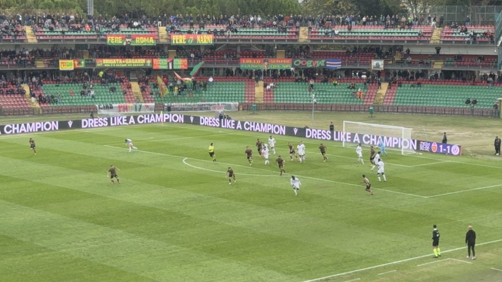 Partita di calcio allo stadio Libero Liberati, con il campo visibile e le squadre in campo. In primo piano una squadra sta attaccando mentre l'altra è in fase difensiva, con alcune persone sugli spalti che osservano l'azione.