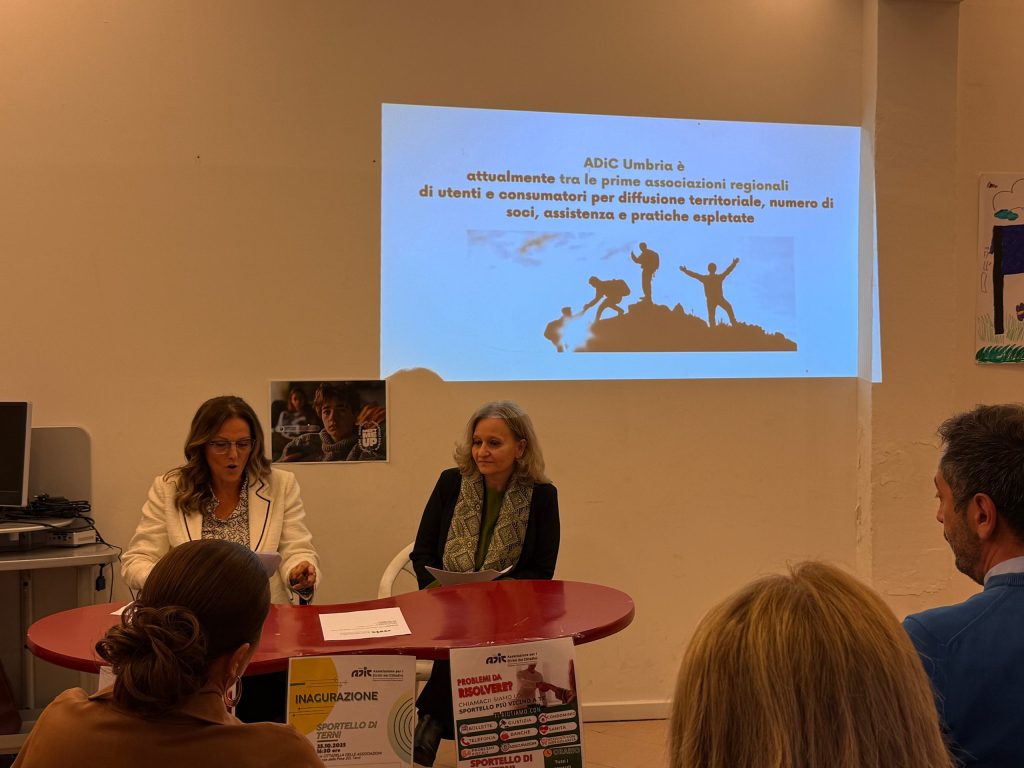 Due donne sedute a un tavolo durante l'inaugurazione dello sportello ADIC, con una presentazione sullo sfondo che evidenzia il ruolo dell'associazione e un'immagine di persone su una montagna.