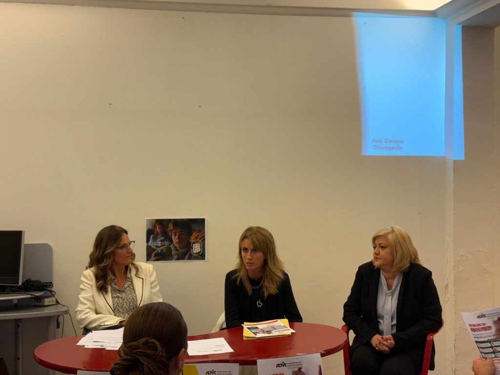 Un gruppo di tre donne sedute attorno a un tavolo rosso durante un evento di inaugurazione, con materiale informativo e un computer visibili sullo sfondo.