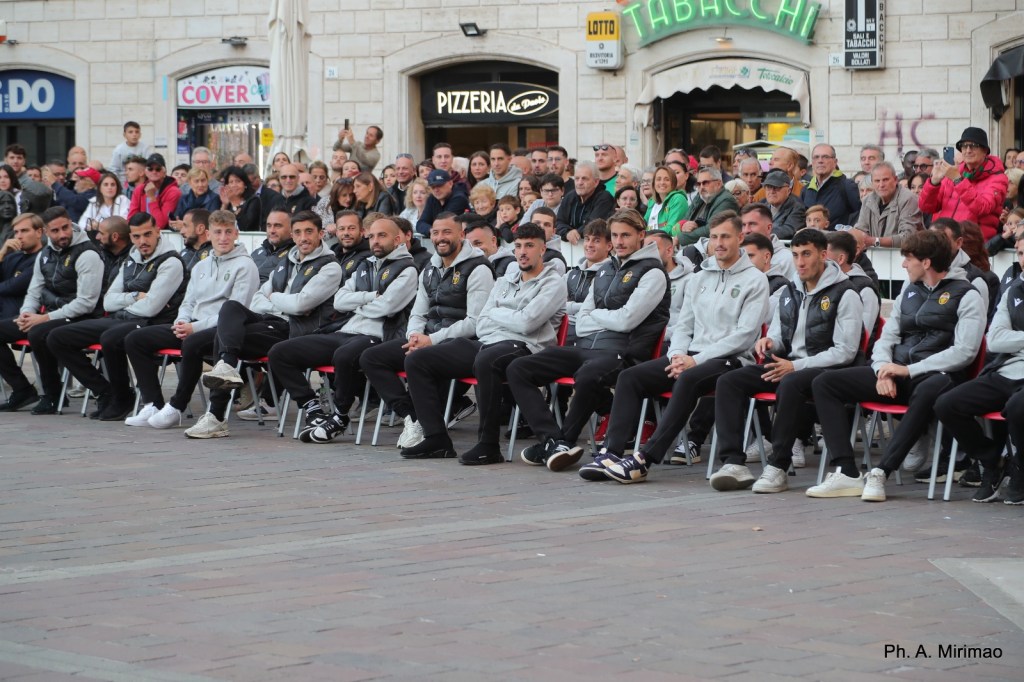 Un gruppo di giocatori della squadra di calcio Ternana seduti in fila durante un evento pubblico, con una folla di tifosi sullo sfondo.