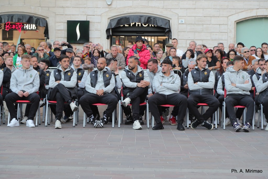 Un gruppo di giocatori della squadra di calcio Ternana seduti su sedie rosse durante un evento pubblico, circondati da una folla di tifosi e sostenitori.