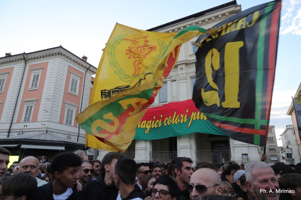 Un gruppo di tifosi in piazza con bandiere gialle e nere, mentre festeggiano la vittoria della squadra di Terni, con un grande striscione sullo sfondo.