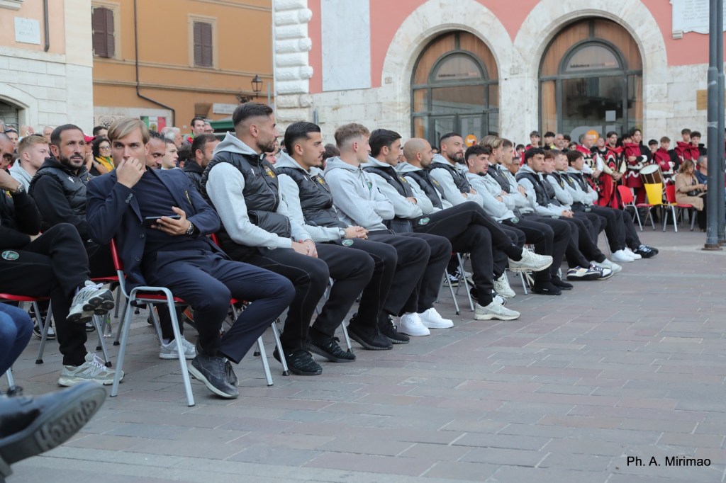 Un gruppo di giocatori seduti su sedie in una piazza, durante un evento pubblico, con alcune persone in costume visibili sullo sfondo.