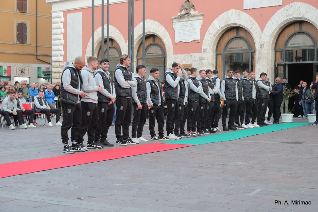 Squadra di calcio della Ternana in piedi su un tappeto rosso durante un evento pubblico in piazza della Repubblica, con il pubblico seduto e in piedi sullo sfondo.