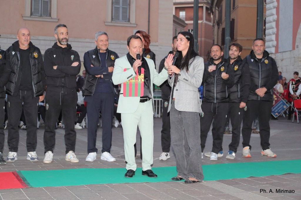 Un momento di interazione durante la festa della squadra di calcio Ternana, con un uomo in abito chiaro che parla a un microfono insieme a una donna in giacca, mentre i membri della squadra sono schierati sullo sfondo.