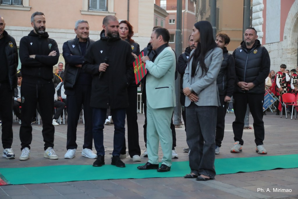 Un gruppo di persone in abbigliamento sportivo si trova in piazza, mentre un uomo in un abito chiaro parla al microfono. Sullo sfondo, sono presenti spettatori e accenni decorativi.