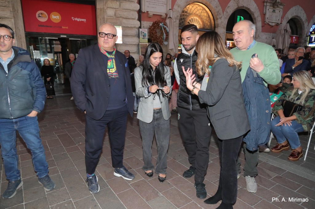 Un gruppo di persone in abiti casual interagisce in una piazza animata, con monumenti e attività che circondano l'area.