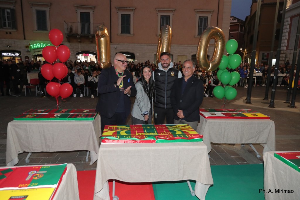 Cerimonia di celebrazione con una torta decorata in piazza, vicino a palloni colorati e una folla di tifosi sullo sfondo.