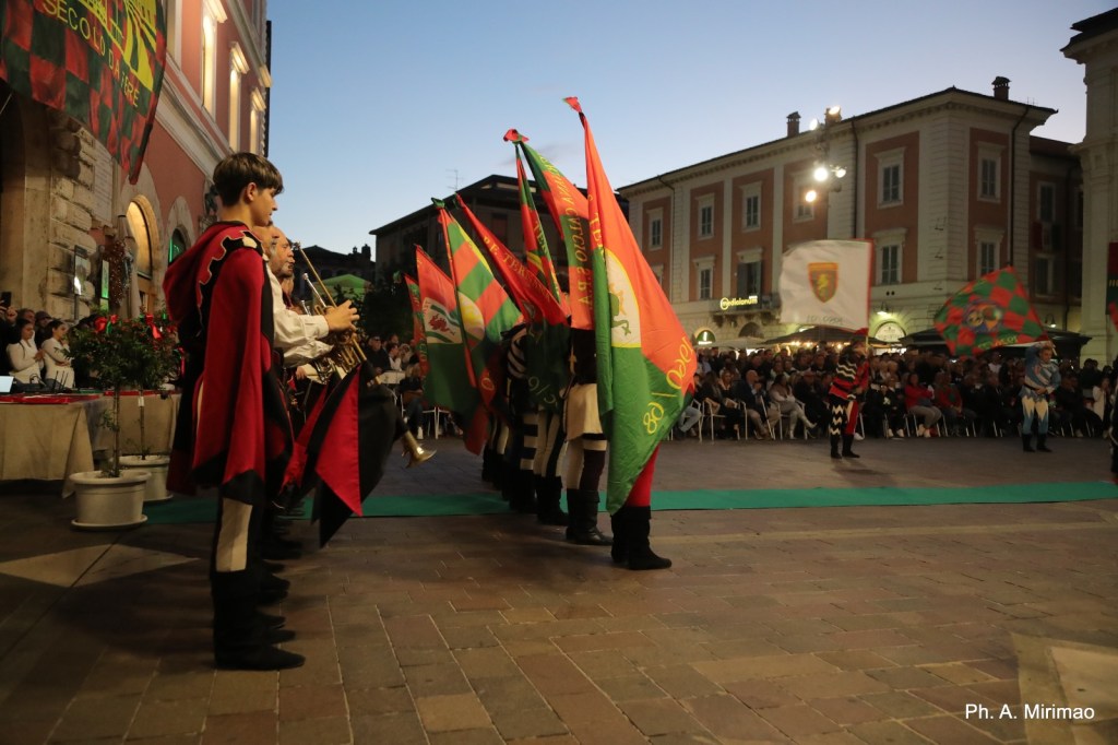 Un gruppo di sbandieratori in costume che esibiscono bandiere colorate in piazza, con una folla in background che assiste all'evento serale.