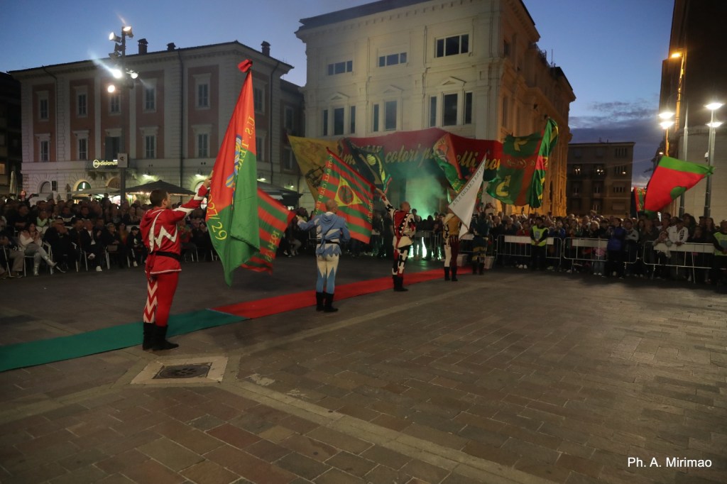 Gruppo di sbandieratori in costume che esibiscono bandiere colorate durante una celebrazione in piazza, con un pubblico presente in sottofondo.