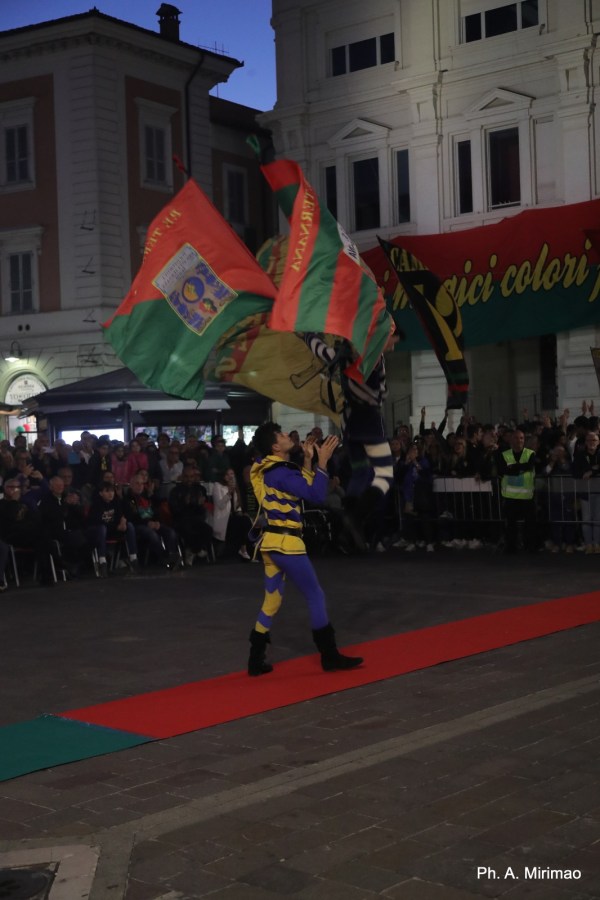 Un performer, vestito in costume colorato, lancia bandiere mentre si svolge una celebrazione in piazza, circondato da un pubblico entusiasta.