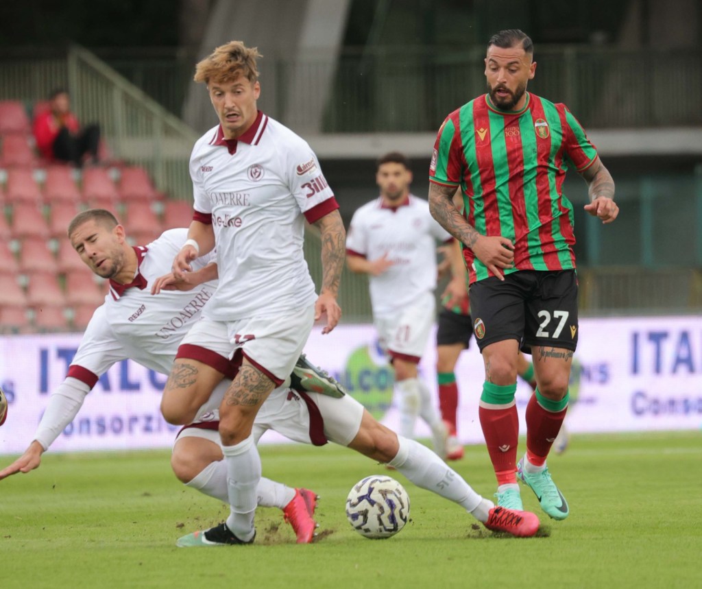 Giocatori di calcio in azione durante una partita, con uno che cade a terra mentre cerca di recuperare il pallone, mentre un avversario si prepara a proseguire il gioco.