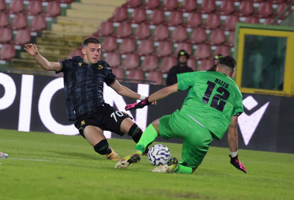 Un giocatore di calcio in maglia scura numero 70 scivola verso un portiere in maglia verde numero 12, mentre quest'ultimo sta bloccando il pallone a terra in un'azione di gioco.