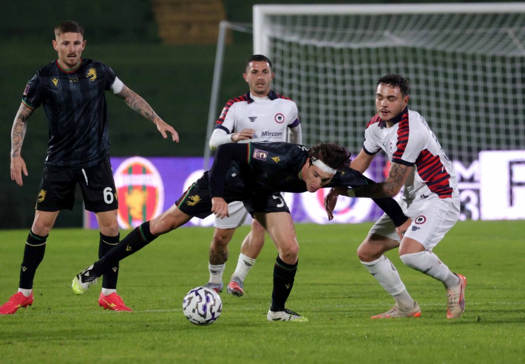 Giocatori di calcio in azione durante una partita, con un atleta in maglia nera che viene placcato da un avversario. Sullo sfondo, altri giocatori si preparano a intervenire.