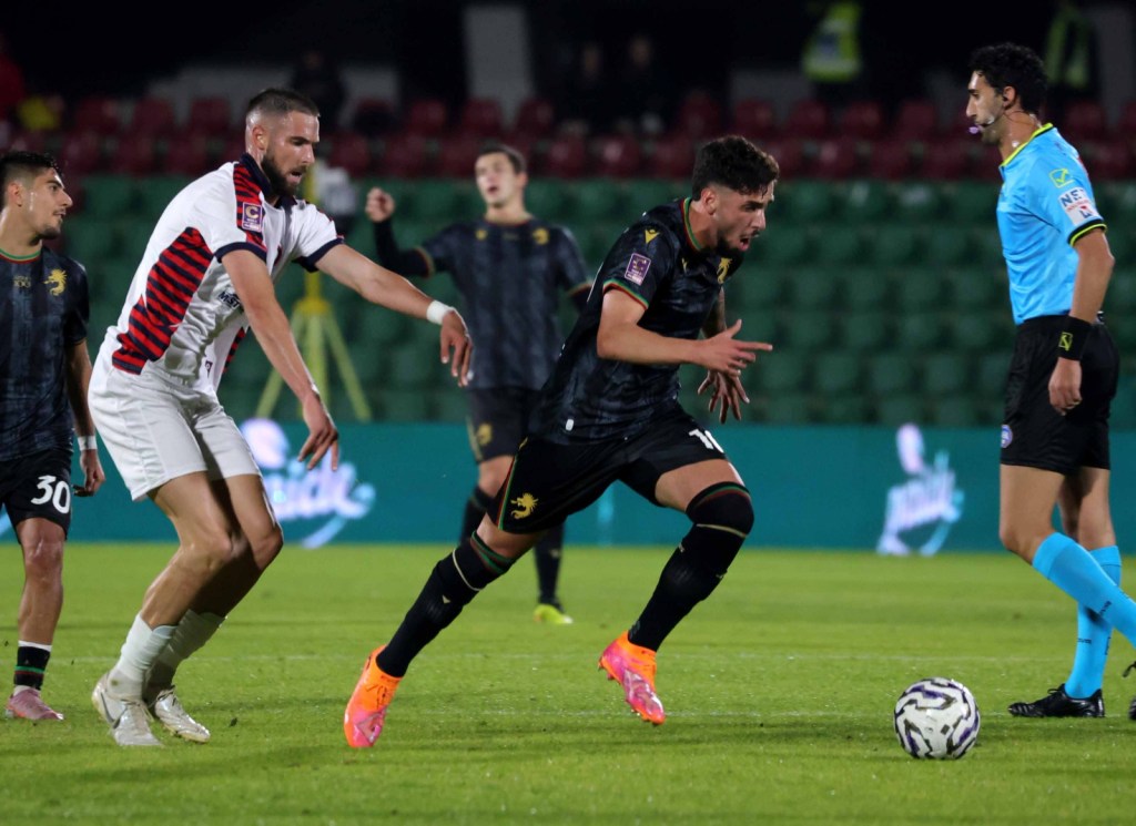 A tense moment in a football match, featuring two players: one in a black kit with bright orange shoes and the other in a white kit with red and black stripes, both vying for the ball.