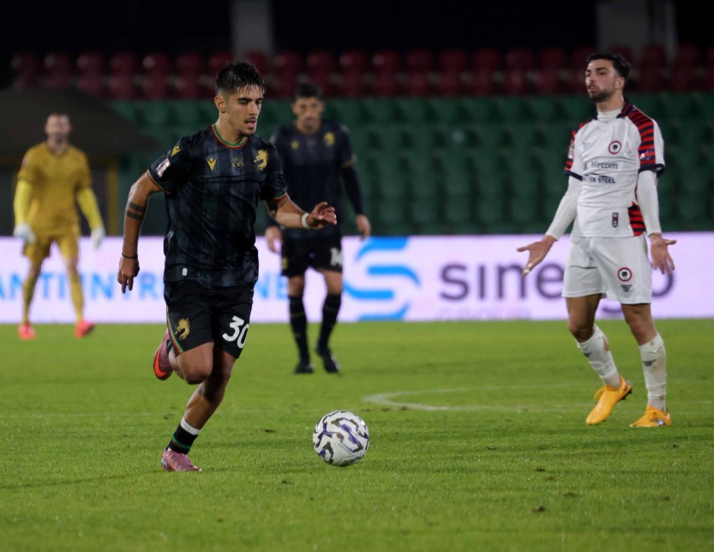 Un giocatore in divisa nera con il numero 30 corre con la palla, mentre un avversario in divisa bianca osserva in distanza. Sullo sfondo, si vede un portiere in divisa gialla.