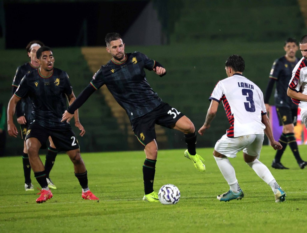 Giocatori in azione sul campo durante una partita di calcio, con un giocatore in maglia scura che tenta di dribblare un avversario in maglia bianca.