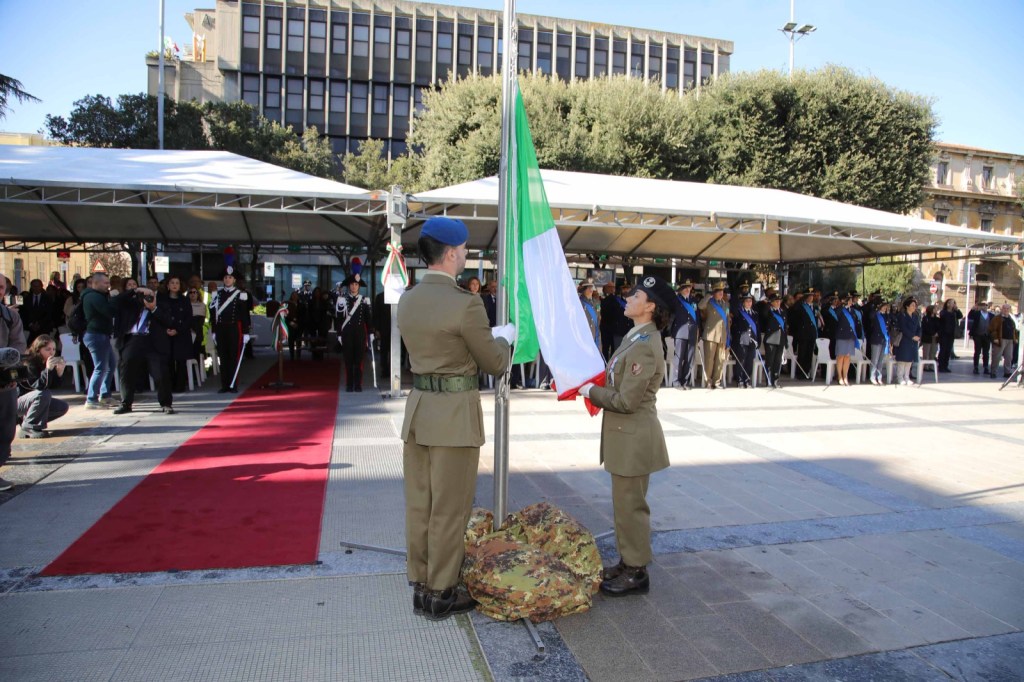 Una donna in abiti formali parla al microfono tenendo un documento, durante una cerimonia commemorativa.