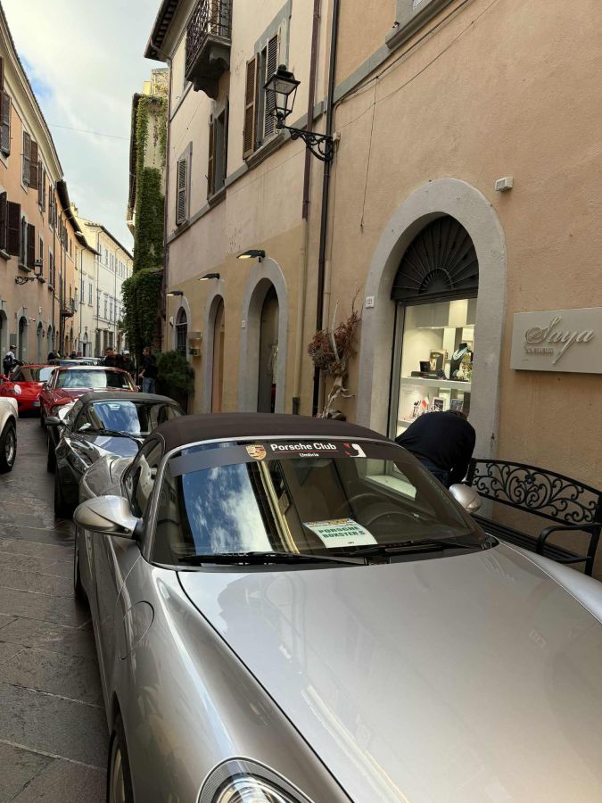 Una via acciottolata con auto d'epoca e supercar, tra cui una Porsche, parcheggiate lungo i lati ed un venditore che interagisce con i passanti.