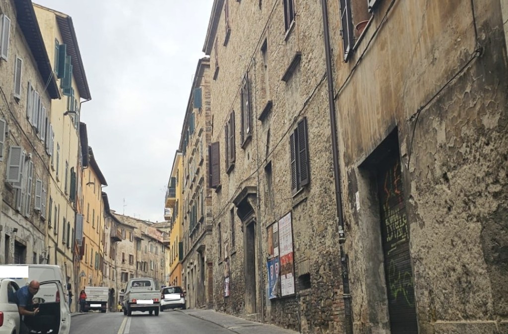A narrow street in Perugia with old stone buildings on either side, cars parked along the road, and a cloudy sky above.