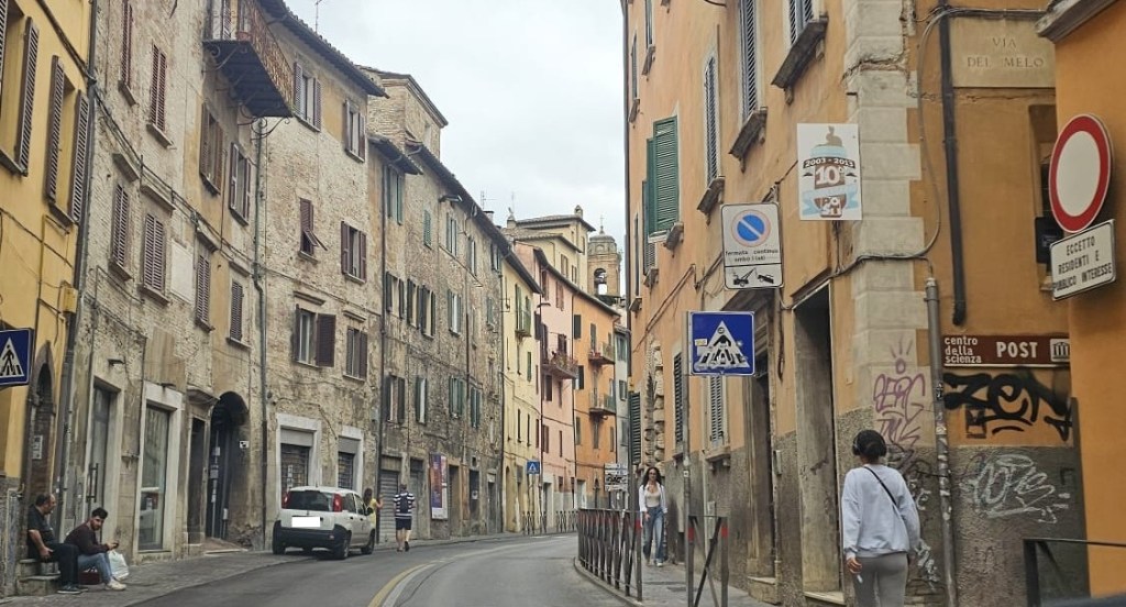 Strada stretta e acciottolata a Perugia, con edifici storici e segnaletica stradale visibile. Una macchina bianca parcheggiata a lato e persone che camminano lungo la via.