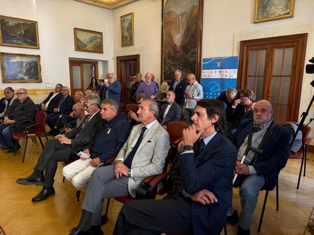 Una sala affollata durante un evento, con molti partecipanti seduti e alcuni in piedi, mentre ascoltano un discorso sul primo memorial D'aloja dedicato alla sostenibilità.