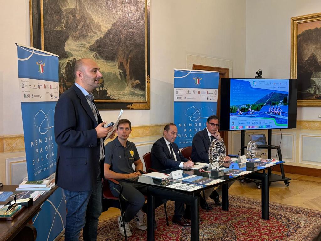 Un relatore presenta il primo memorial D'aloja dedicato alla sostenibilità, con partecipanti seduti a un tavolo, un display alle spalle e opere artistiche sul muro.