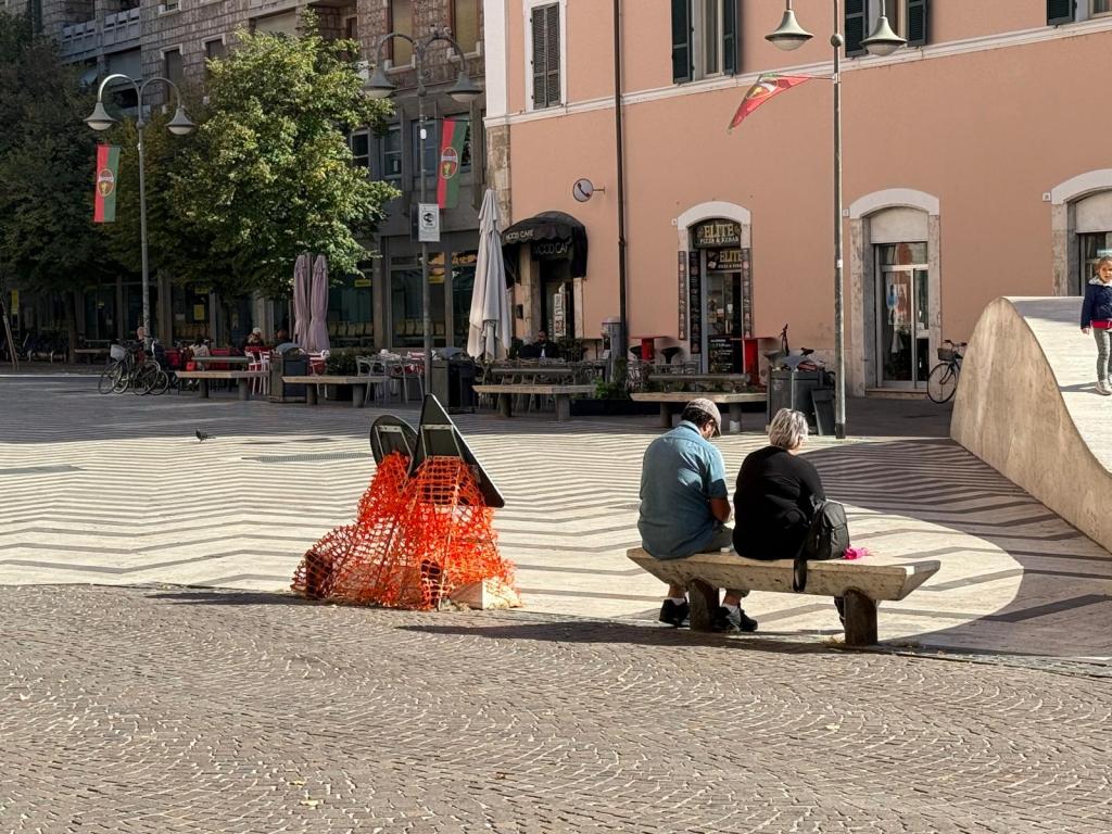 Two people sitting on a bench in a public plaza, surrounded by trees and buildings. One person has short hair and is wearing a blue shirt, while the other has grey hair and is wearing a black shirt. In the foreground, there is a construction barrier and cobblestone pavement.