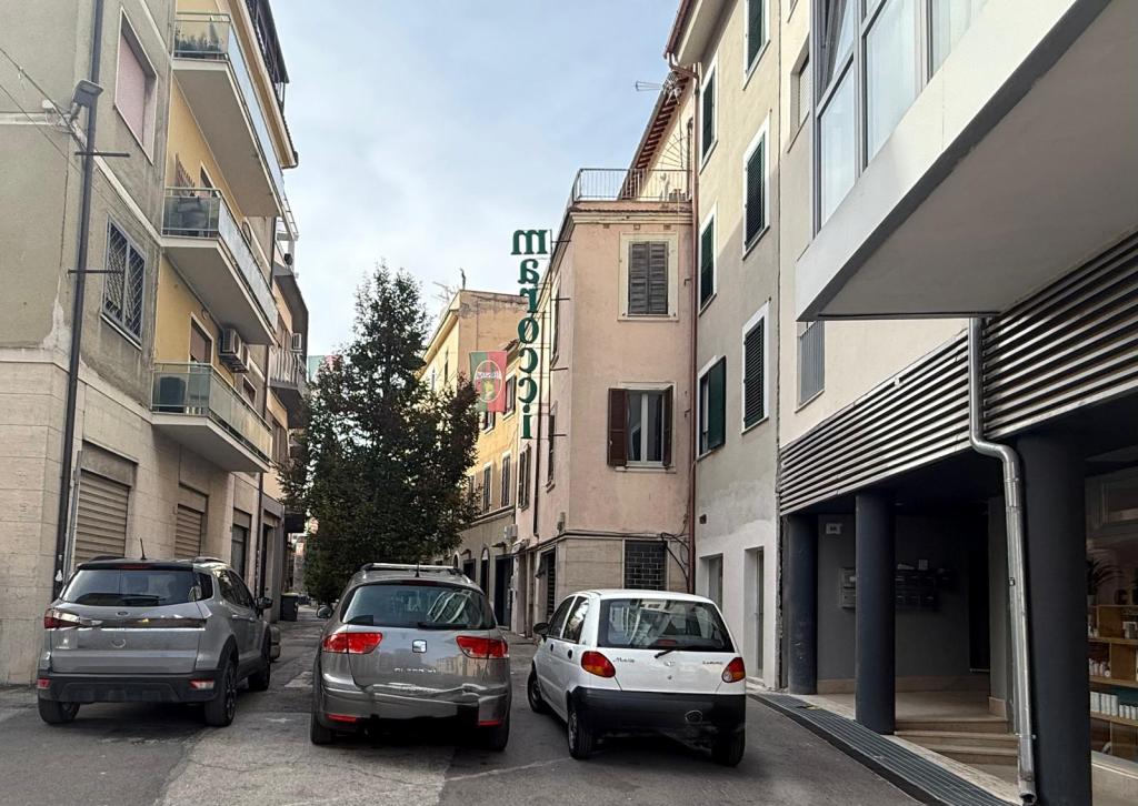 Narrow street view featuring parked cars on both sides and buildings with balconies, including a sign in green letters.