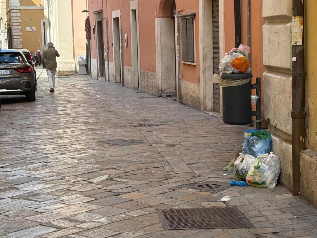 A narrow street lined with buildings. A person walks away in the distance. Trash bags are placed next to a garbage bin on the sidewalk.