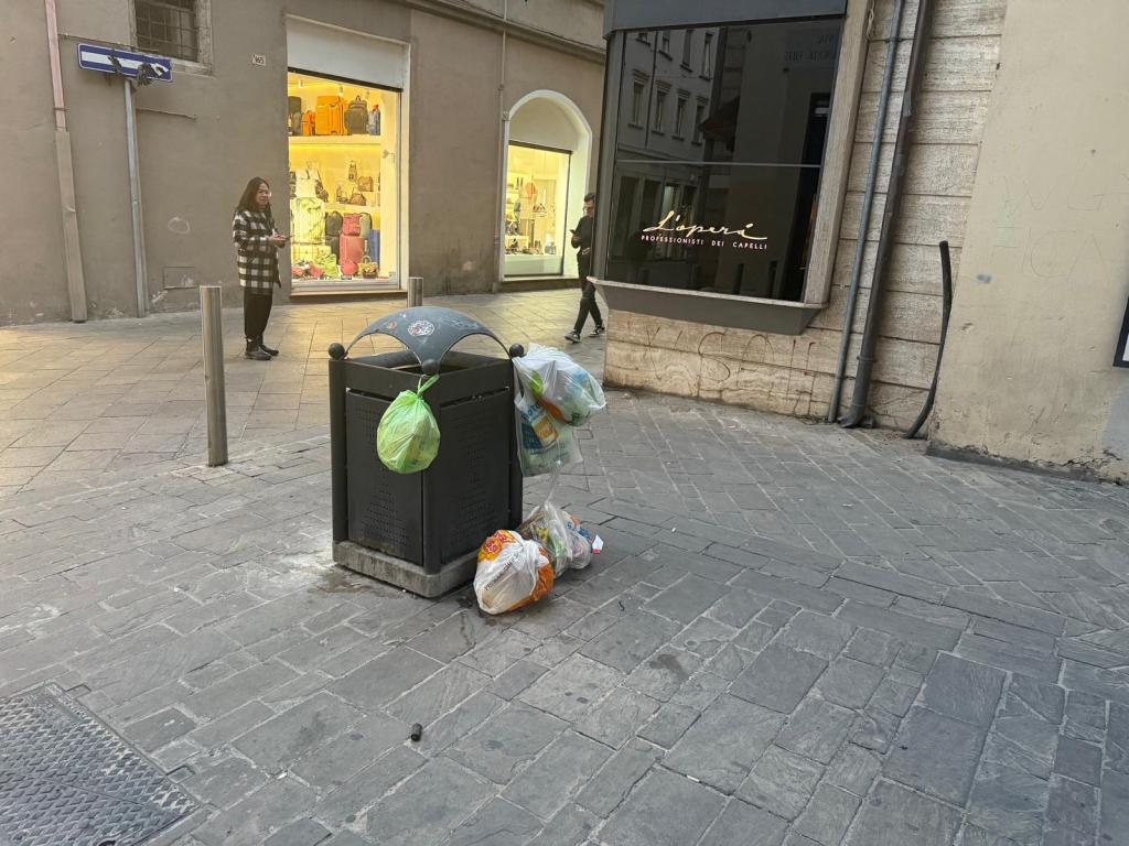 A public trash can overflowing with garbage on a city street, with a passerby in the foreground and a store display visible in the background.