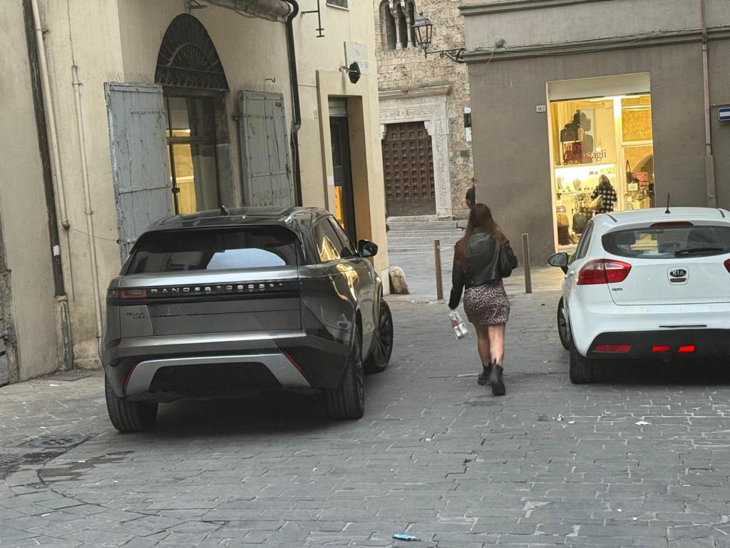 A woman walking down a narrow street past two parked cars, one is an electric SUV and the other a white compact car. In the background, there are shops and historic architecture.