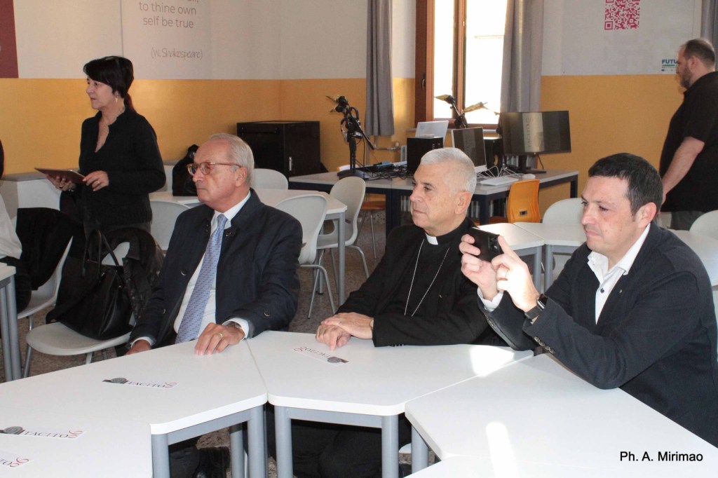 A group of three men seated at tables in a brightly lit room, with a woman standing in the background holding a tablet. The first man on the left is in a suit, the middle man is wearing clerical attire, and the third man is dressed casually while holding a camera.