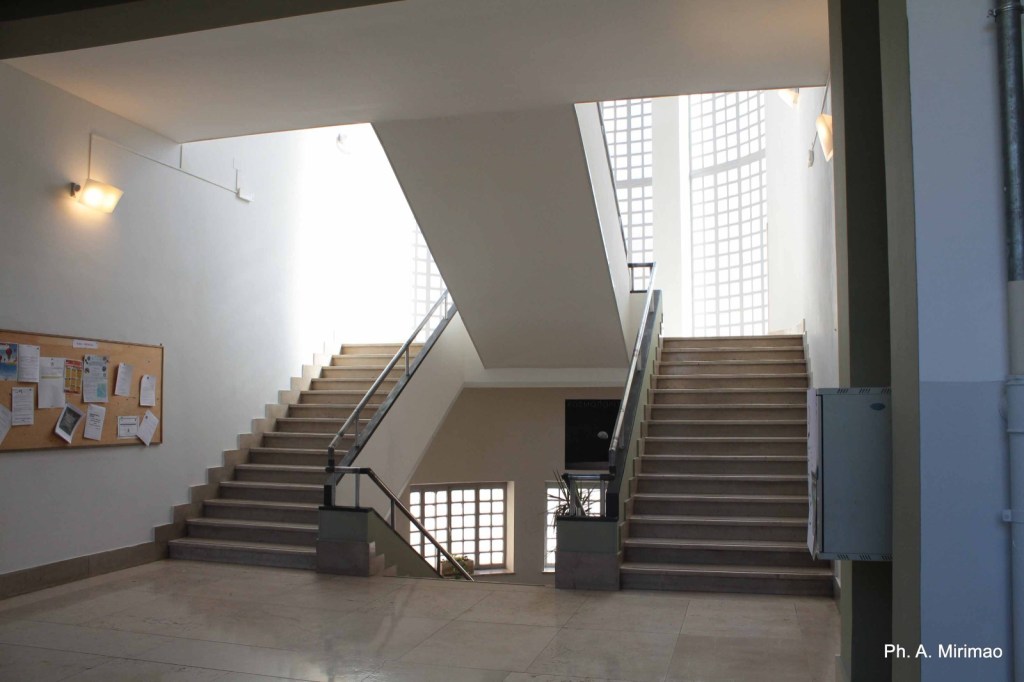 A well-lit staircase with two sets of stairs leading up, surrounded by white walls and a bulletin board filled with papers.