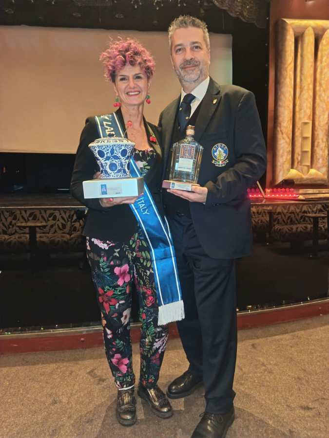 A woman with short, curly pink hair and a floral outfit stands next to a man in a suit. She holds a decorative vase and a trophy, while he presents a bottle. Both are smiling in a theater setting.